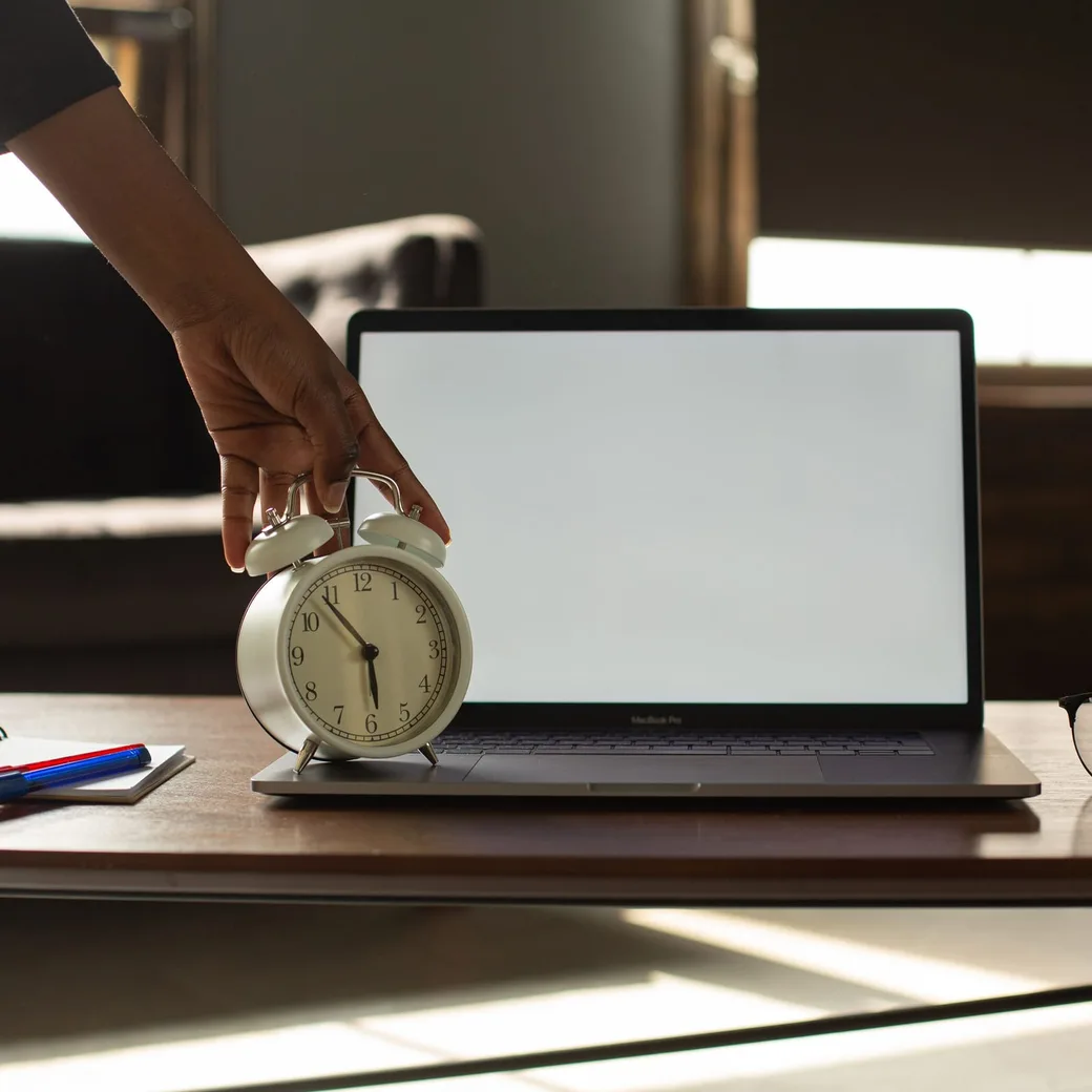 Una mano deja un despertador sobre la mesa, junto a un portátil